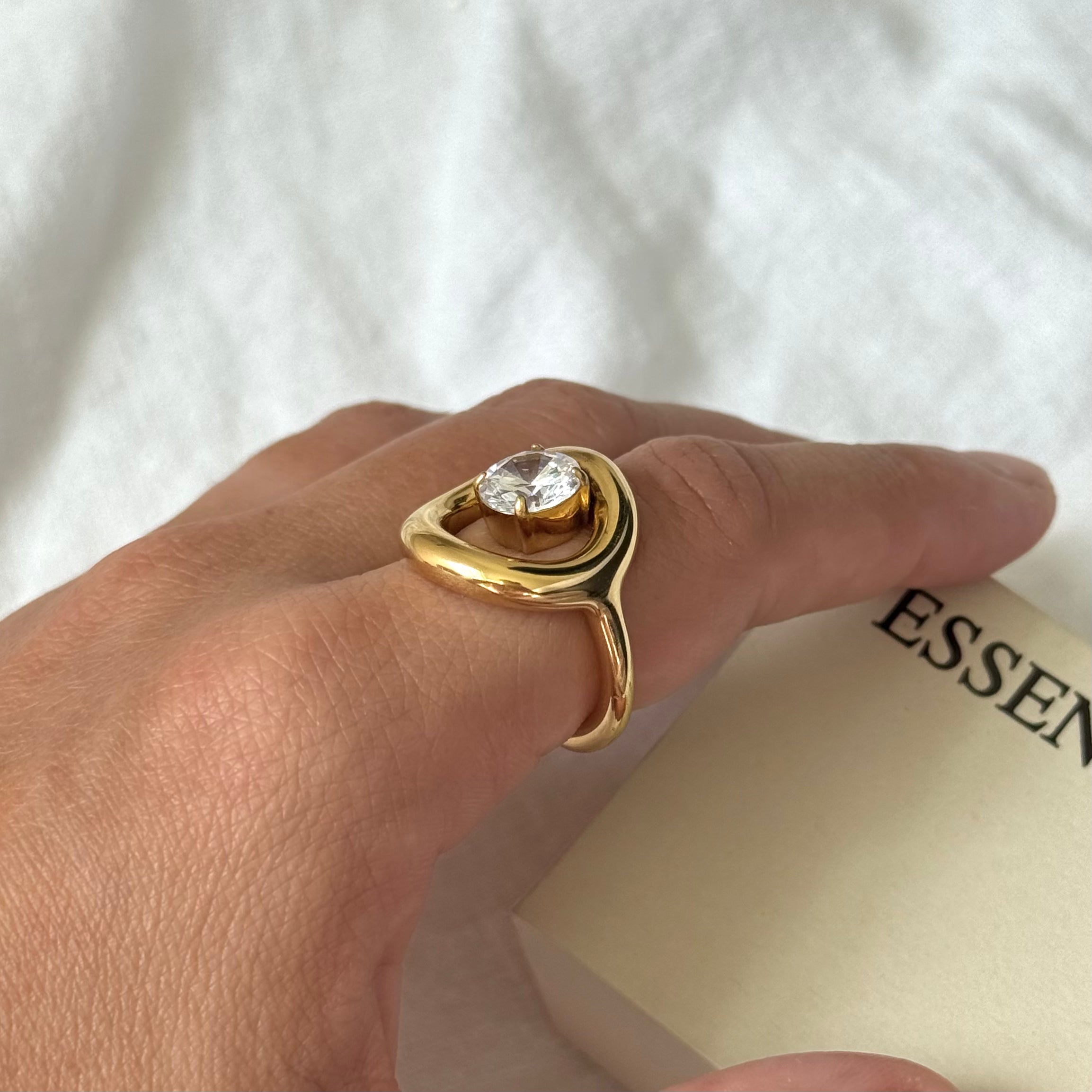 A gold Orbit Ring with a clear round zircon, worn on a person's finger against a soft white fabric background with a beige card partially visible.