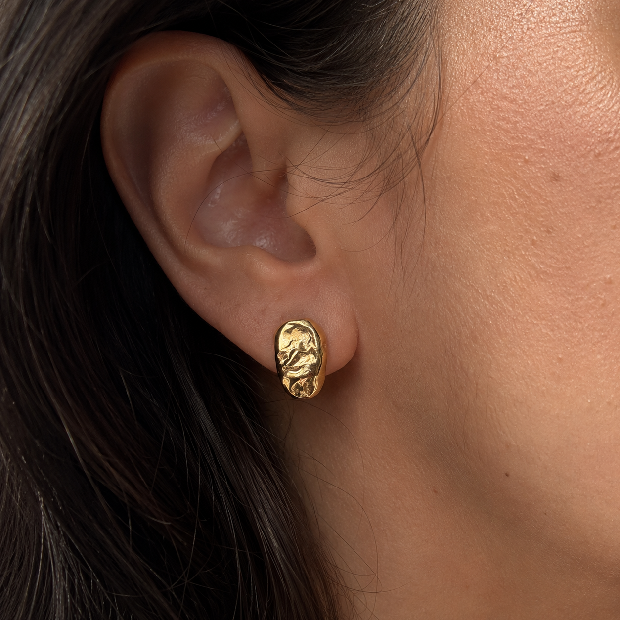 A close-up of a person's ear wearing a small molten organic gold stud earring.
