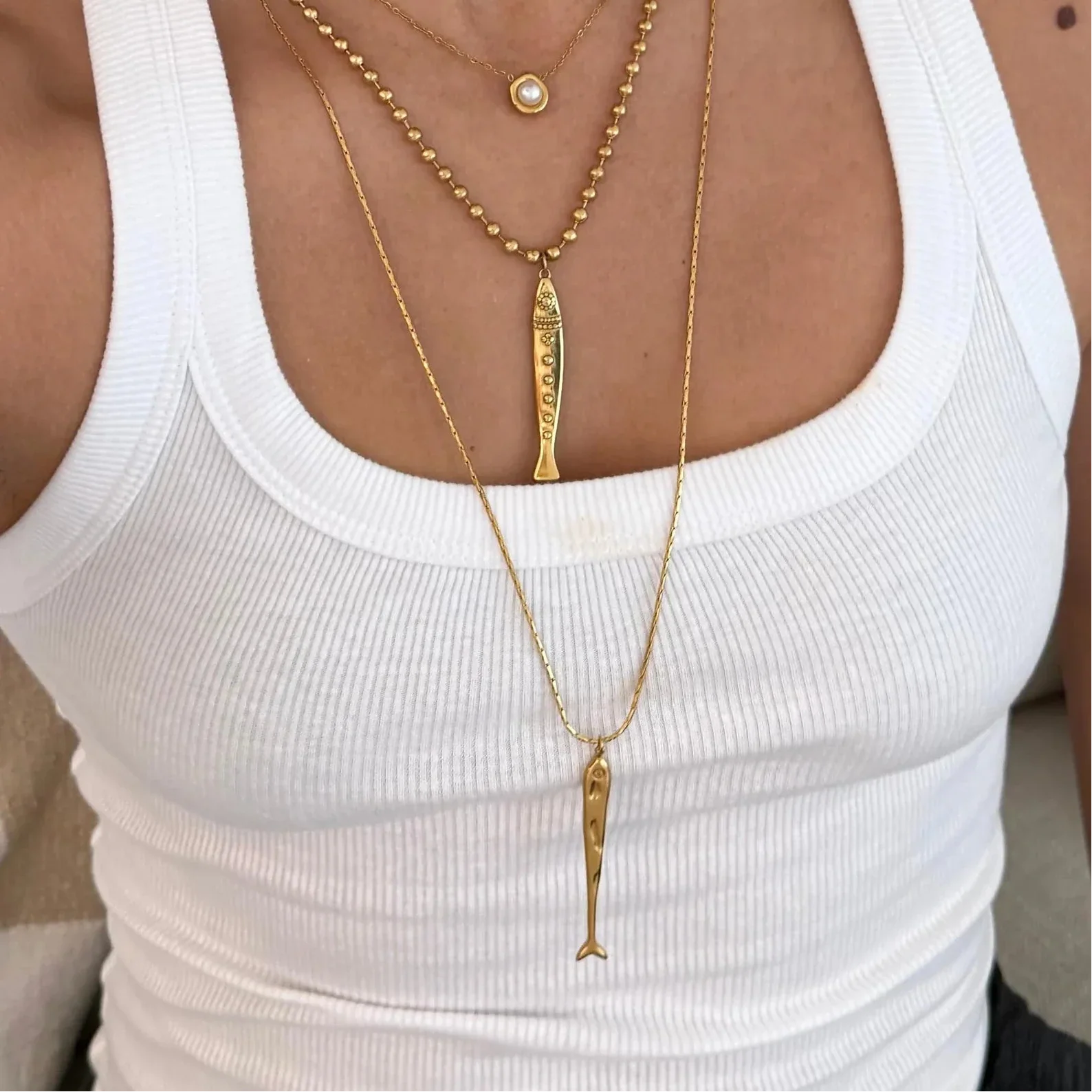 A close-up of a person wearing layered gold necklaces over a white ribbed tank top. The necklaces include a short beaded chain with a small round pendant, a longer chain with a vertical bar pendant, and a long chain with a fish-shaped pendant.