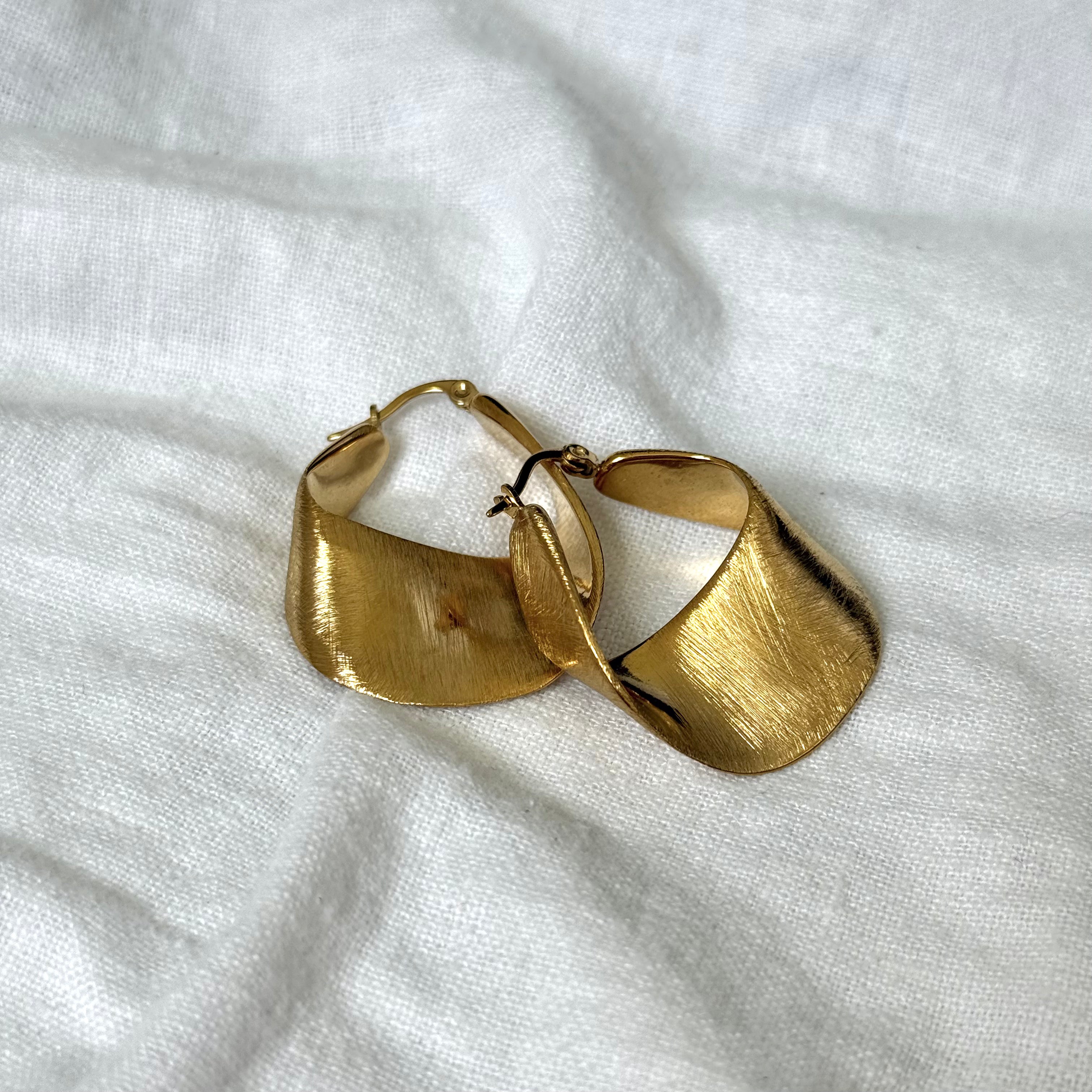 A pair of textured Wavy Hoop Earrings with a wide, curved, leaf-like shape, displayed on a white linen fabric background.
