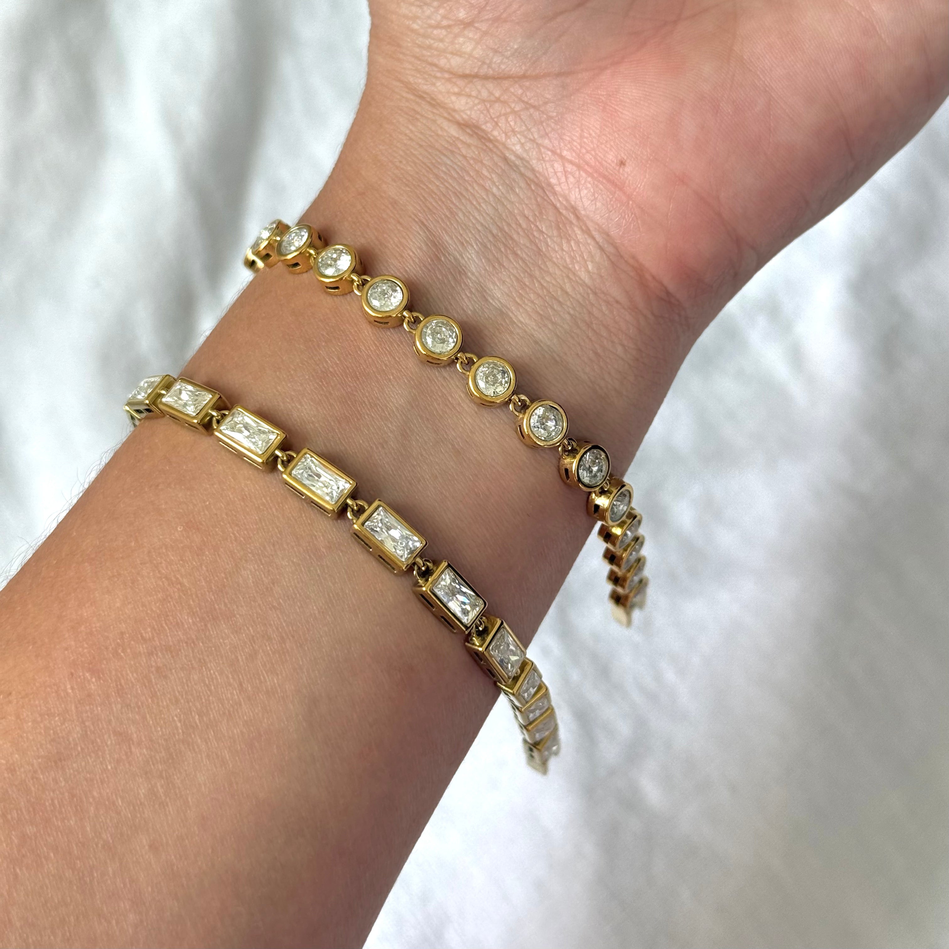 Two gold tennis bracelets with clear zircon bezel stones worn on a person's wrist against a white fabric background.