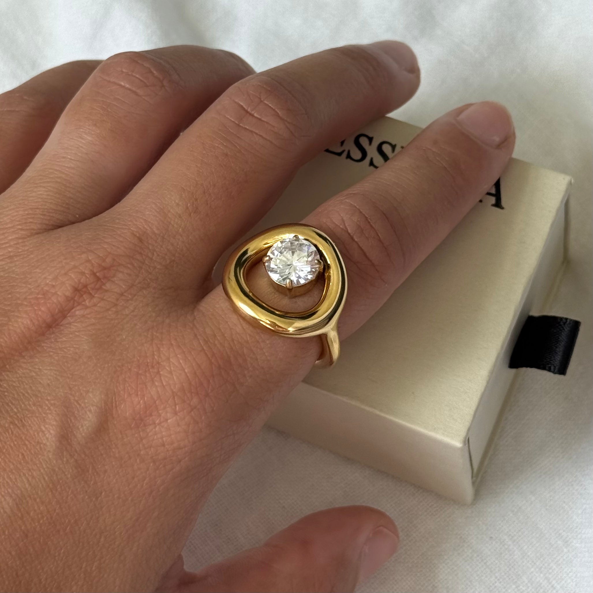 A gold ring with a round clear zircon is worn on a person's finger, positioned over a cream-colored jewelry box.