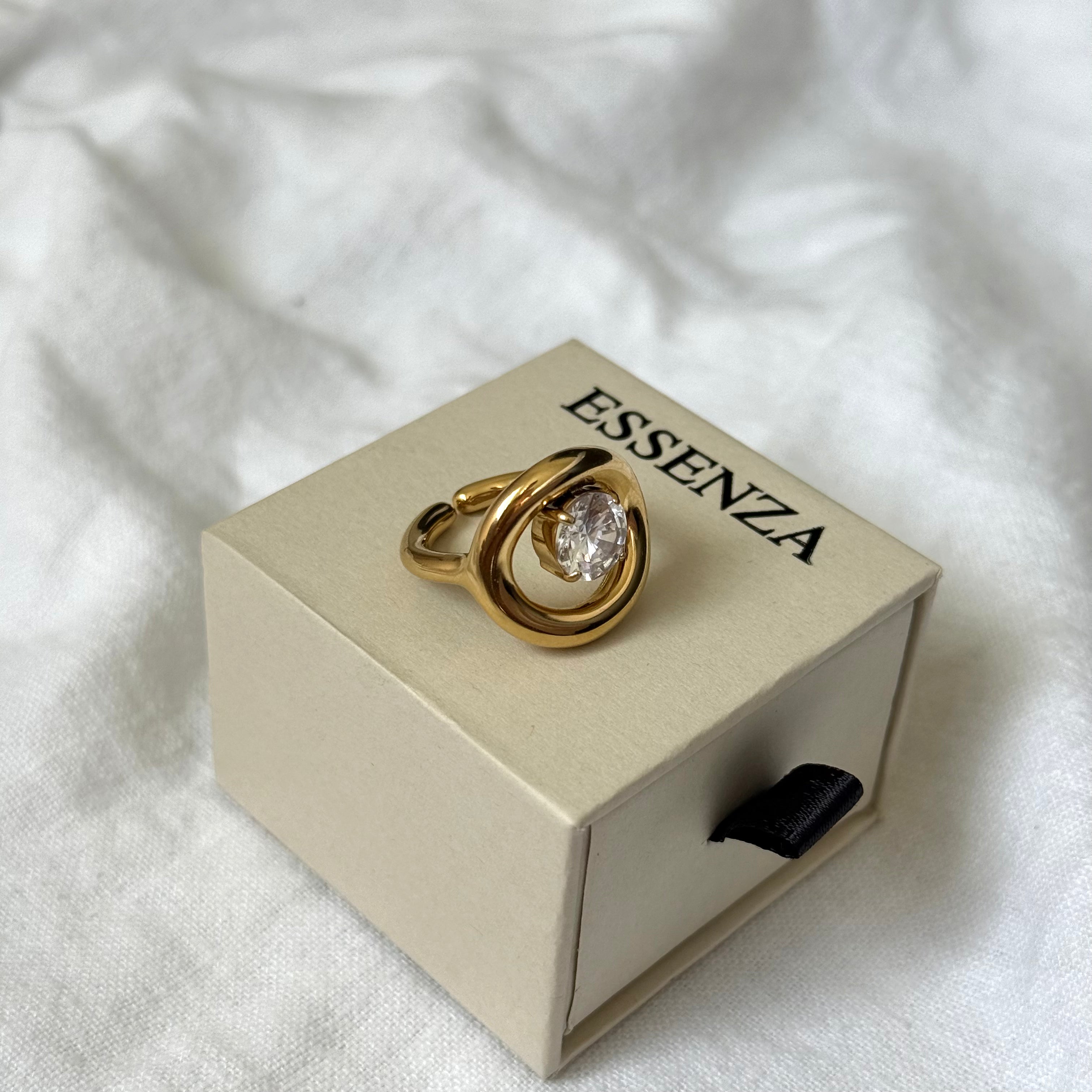 A gold Orbit Ring with a clear round zircon resting on a beige Esenza jewelry box, set against a soft white fabric background.