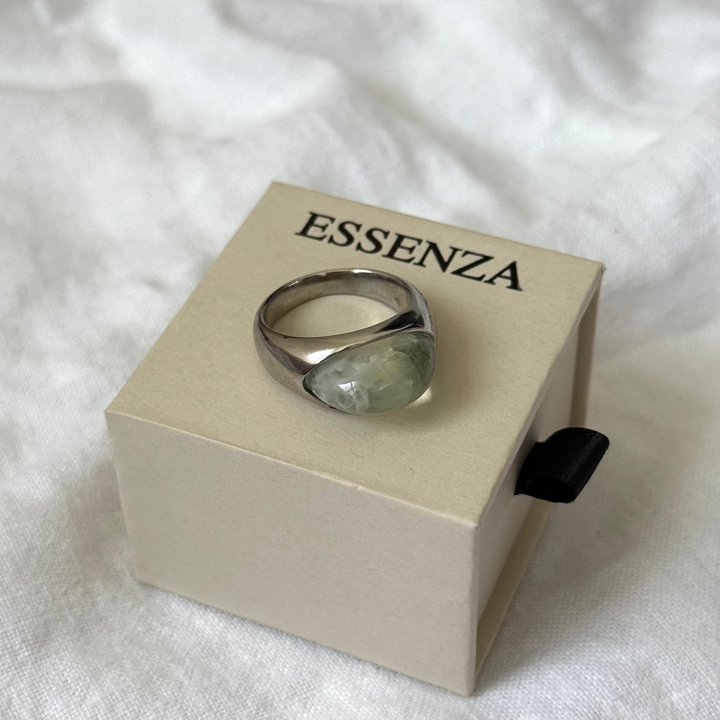 A silver ring with a large, irregular light green stone, displayed on a beige Esenza jewelry box over a white fabric background.