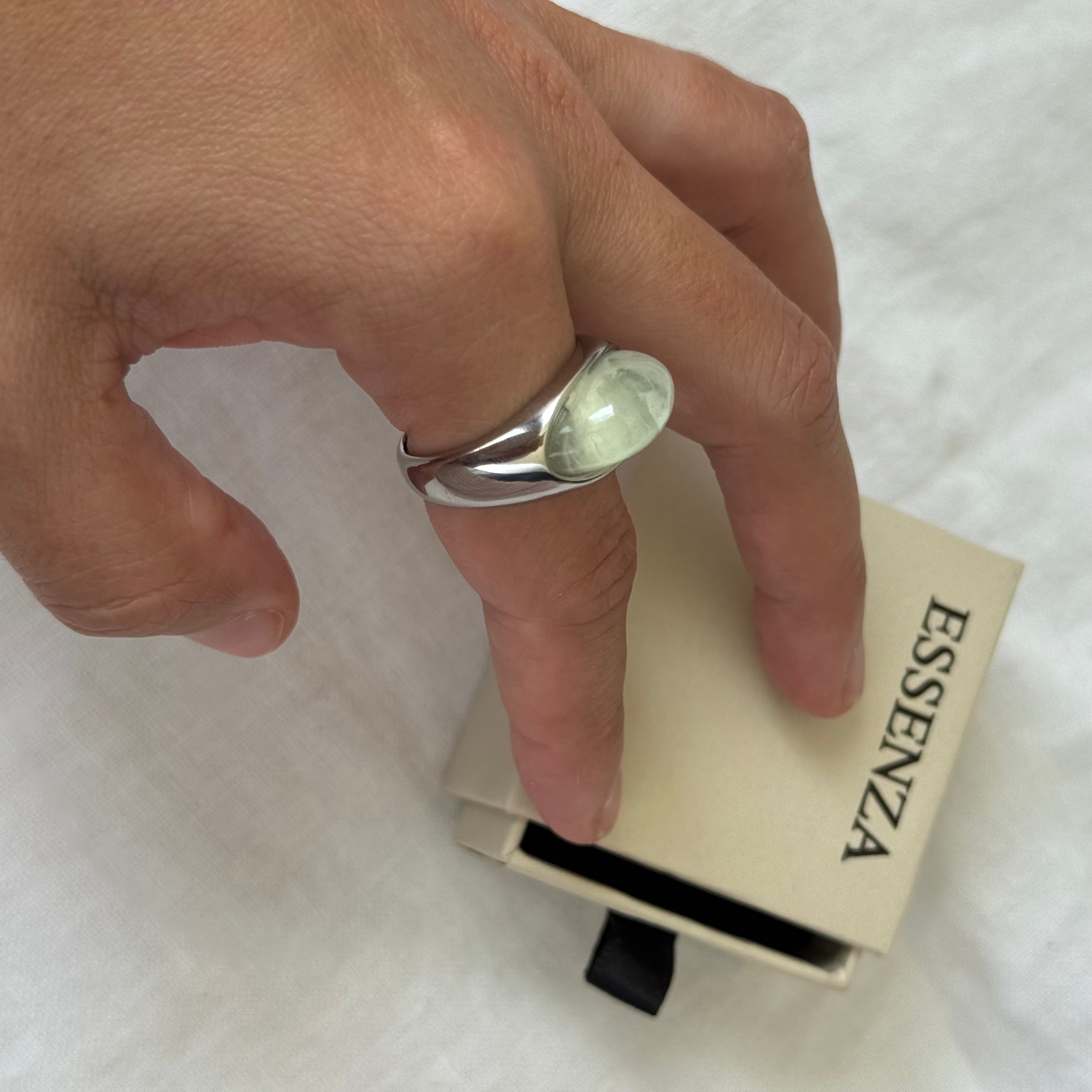 A hand wearing a silver-toned ring with a large oval pale green stone, resting near an open cream-colored ESSENZA jewelry box on a white fabric background.