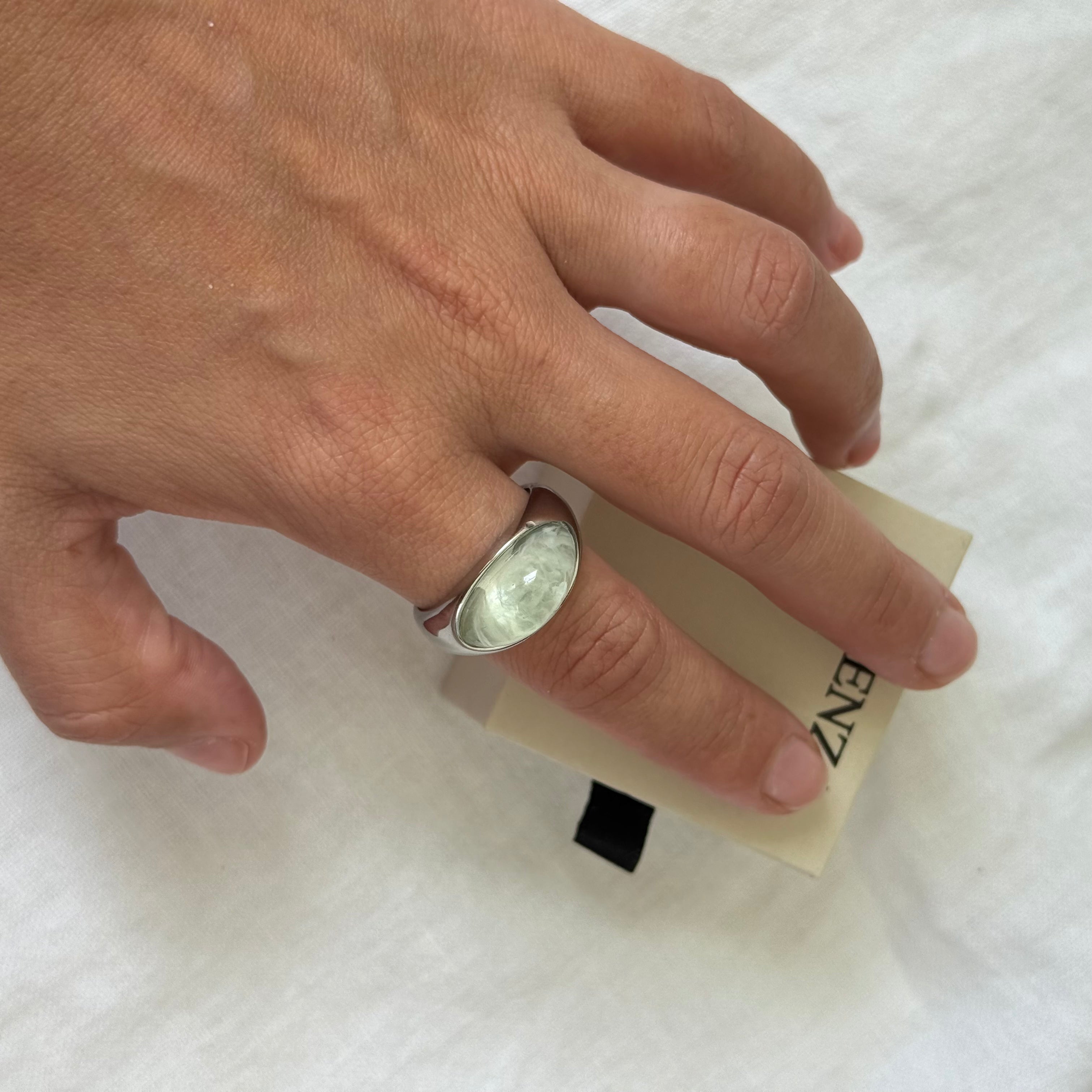 A silver ring with a round, polished clear stone worn on a person's ring finger, resting on a beige paper tag with the brand name 'ENZ' partially visible, set against a white fabric background.