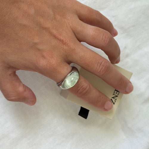 A silver ring with a round, polished clear stone worn on a person's ring finger, resting on a beige paper tag with the brand name 'ENZ' partially visible, set against a white fabric background.