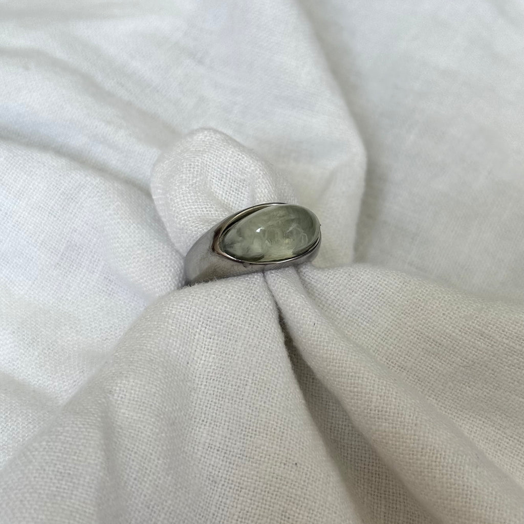 A silver ring with an oval, polished gray-green stone, displayed on a textured white fabric ring stand.