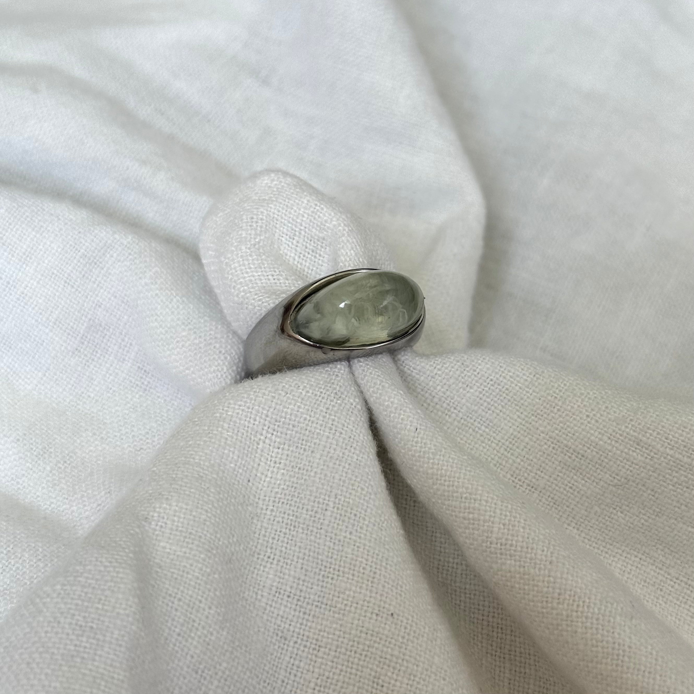 A silver ring with an oval, polished gray-green stone, displayed on a textured white fabric ring stand.