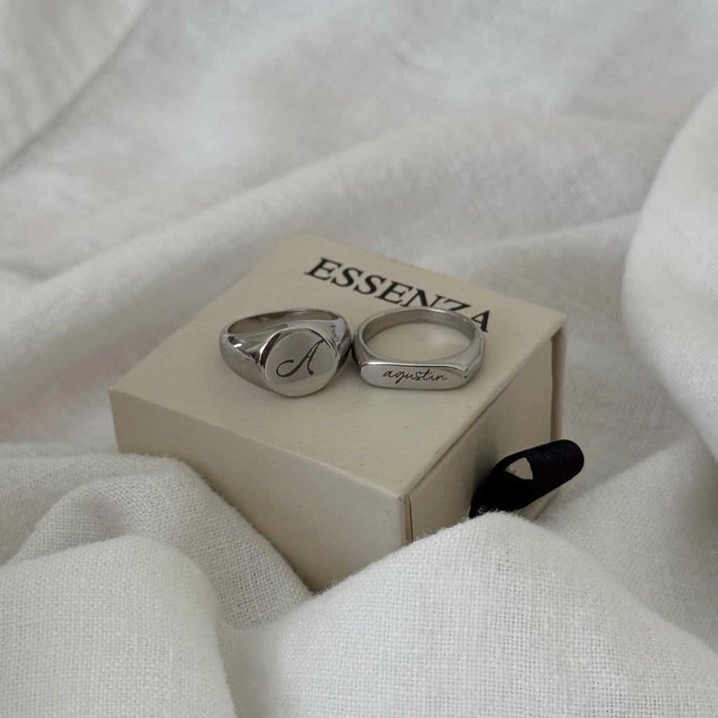 Two engraved silver rings resting on a beige ESSENZA box, surrounded by soft white fabric.