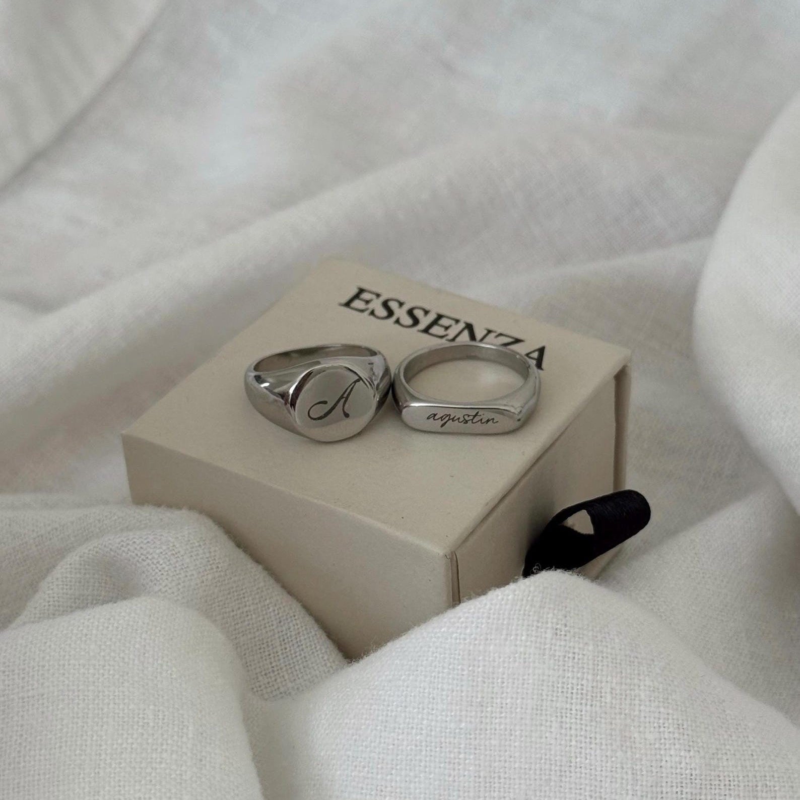 Two engraved silver rings resting on a beige ESSENZA box, surrounded by soft white fabric.