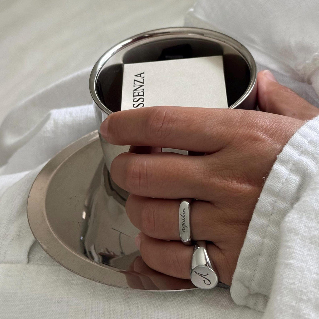 A close-up of a hand wearing a silver ring with a name engraved, holding a small silver tin labeled SSENZA, resting on a white textured blanket.