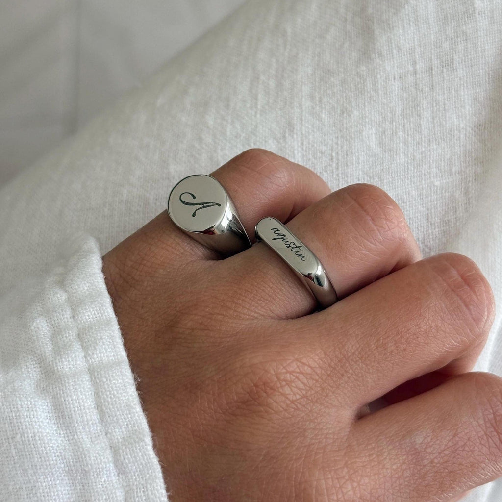 A close-up of a hand wearing a silver signet ring engraved with the initial "A" against a soft white fabric background.