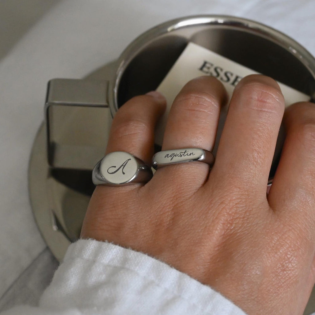 A close-up of a person's hand wearing two silver rings: a round signet ring with an engraved letter 'A' on a white face, and a slim band ring engraved with the name 'Agustin'. The hand is resting near a metallic cup.
