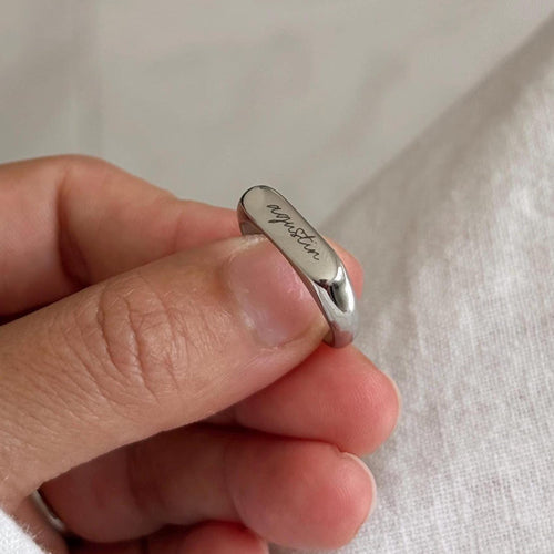 A close-up of a hand holding a polished silver band ring engraved with the name 'Agustin' against a soft, light-colored fabric background.