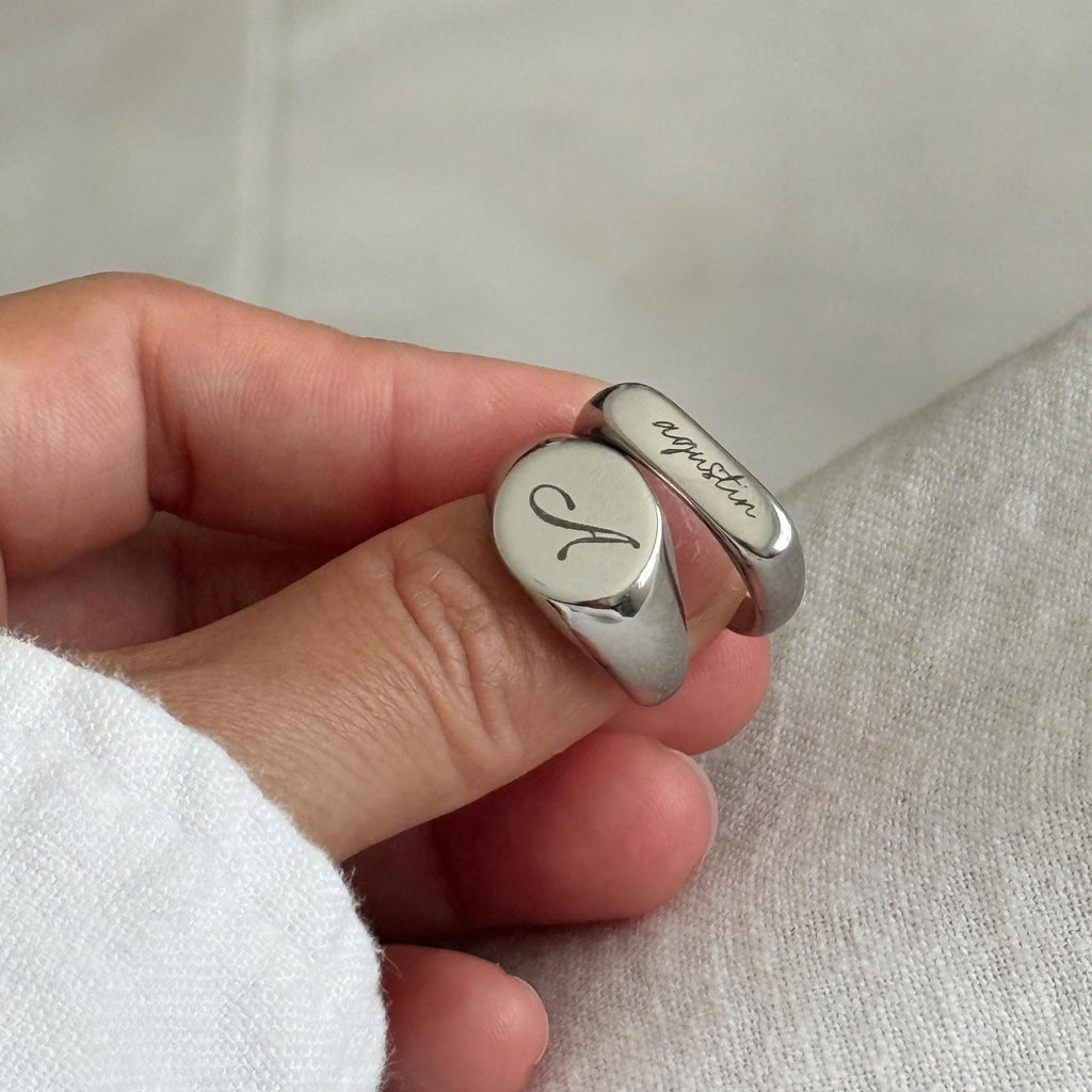 A close-up of a hand holding a polished silver signet ring engraved with a cursive 'A' on one side and the name 'Agustin' on the other, set against a neutral textured fabric background.