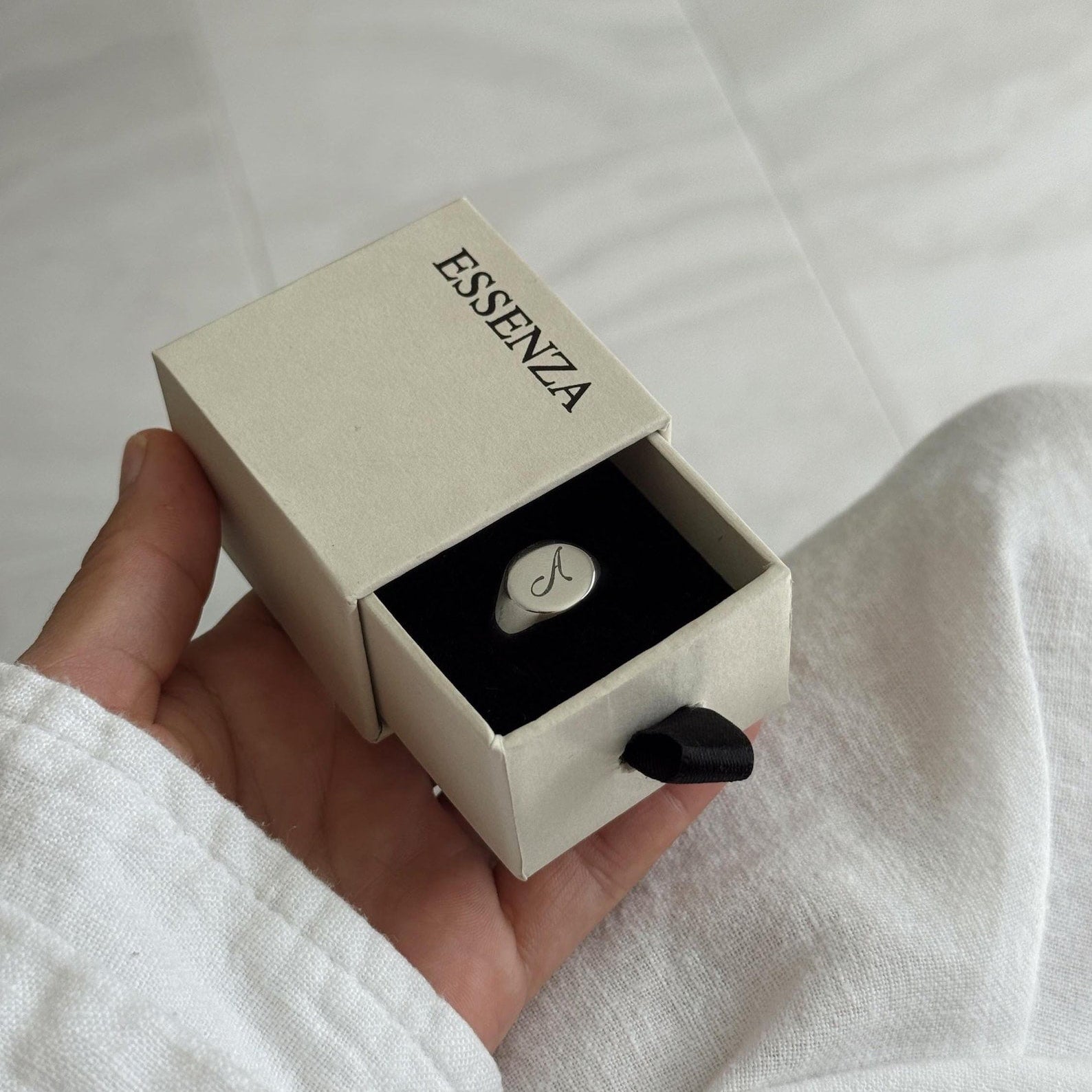 A hand holding an open beige ESSENZA jewelry box that reveals a silver signet ring with an engravedl 'A' on a white textured fabric background.