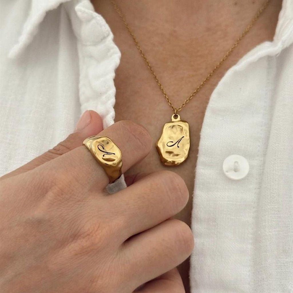 A close-up lifestyle shot of a person wearing a organic shaped gold ring and matching gold pendant necklace, both engraved with the letter 'A', styled with a white button-down shirt.
