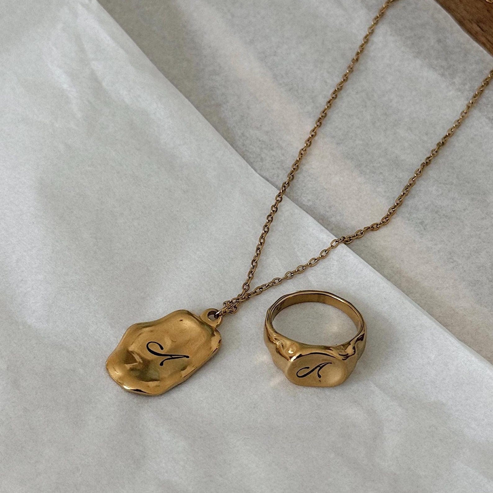A gold necklace with a textured, irregularly shaped pendant and a matching gold ring, both engraved with the cursive letter 'A', displayed on a white textured paper background.