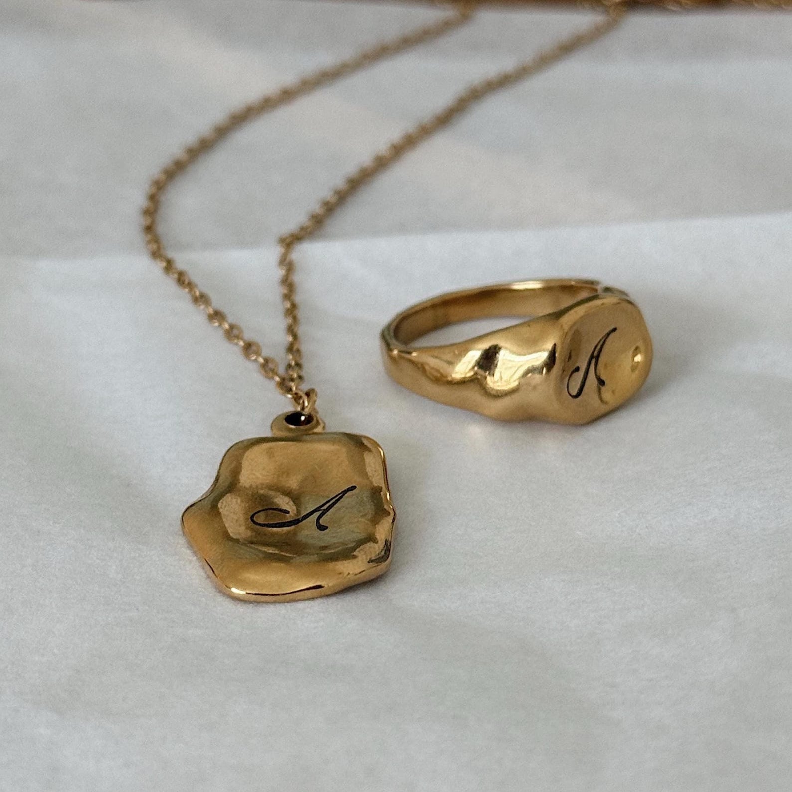 A gold necklace with a wavy, irregular pendant engraved with a cursive letter 'A', displayed next to a matching gold signet ring with the same letter on a white textured surface.