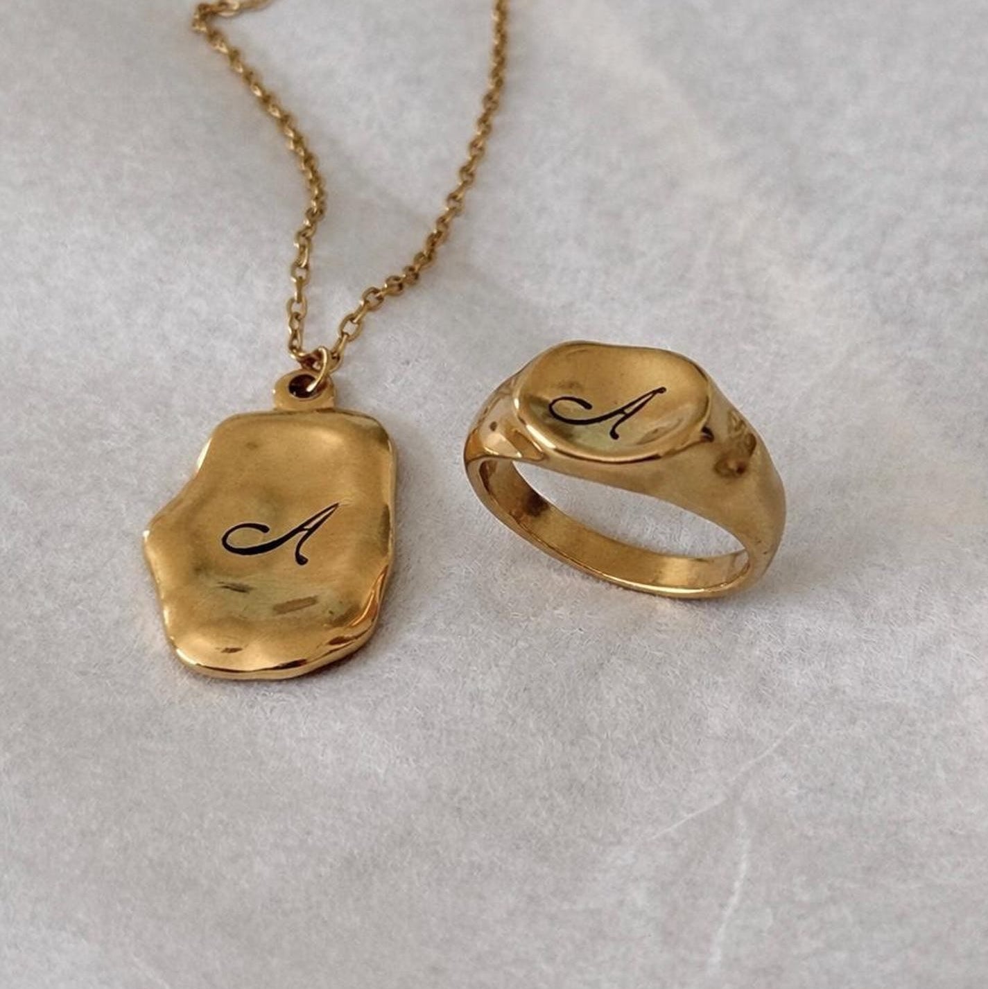A gold necklace and matching ring with a polished, irregularly shaped pendant engraved with the letter 'A', displayed on a textured light gray surface.