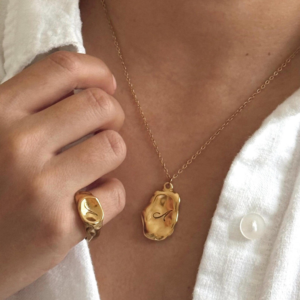A close-up lifestyle shot of a person wearing a gold Pebble Initial Necklace engraved with the letter 'A', along with a matching gold ring on their finger, styled with a white linen shirt.