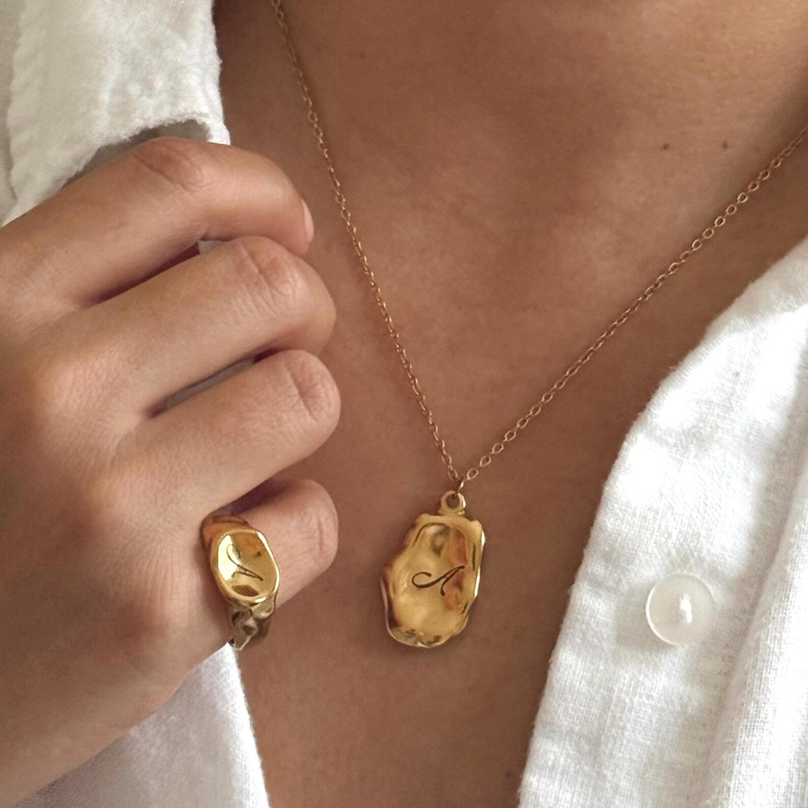 A close-up lifestyle shot of a person wearing a gold Pebble Initial Necklace engraved with the letter 'A', along with a matching gold ring on their finger, styled with a white linen shirt.