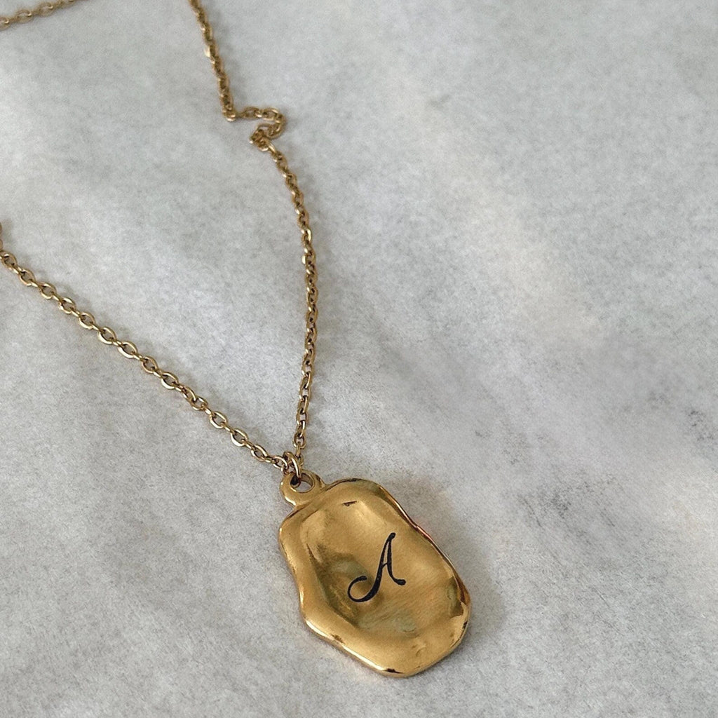 A gold necklace with an irregular, polished pendant engraved with a black cursive letter 'A', displayed on a textured light gray surface.