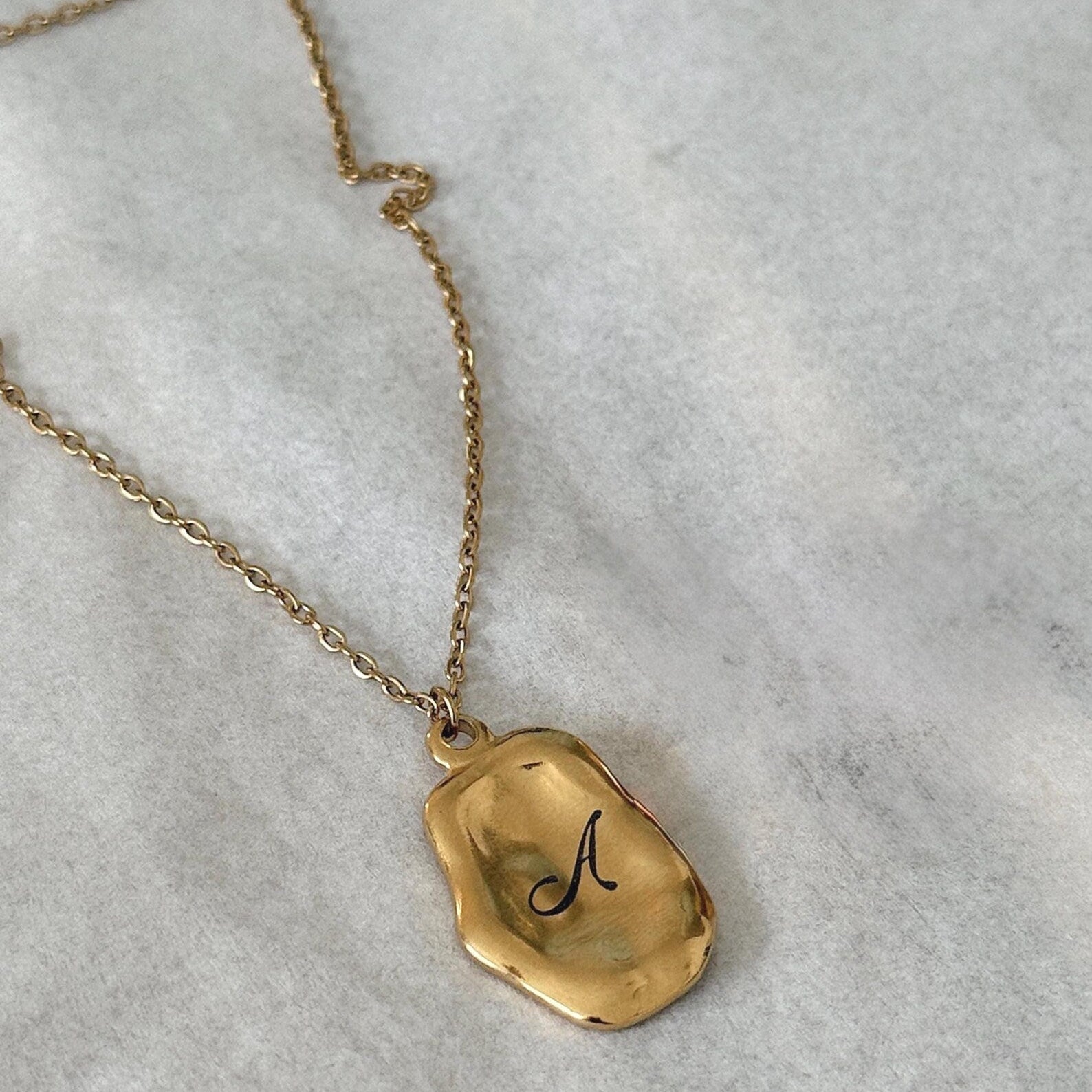 A gold necklace with an irregular, polished pendant engraved with a black cursive letter 'A', displayed on a textured light gray surface.