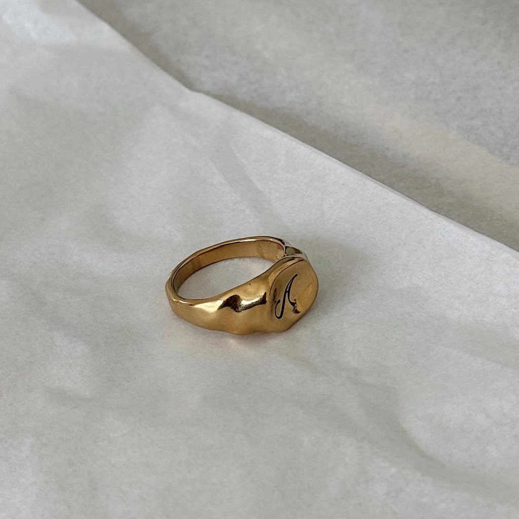 A polished gold organic shaped signet ring with a custom engraved cursive monogram, resting on a textured white surface.
