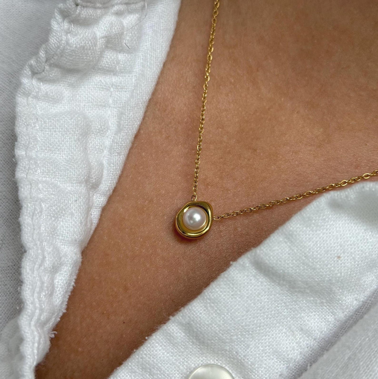 A close-up of a gold chain necklace with a small oval gold pendant holding a white pearl, worn on a person's neck over a white textured top.