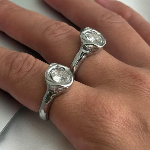 A close-up of a hand wearing two silver molten rings with bezel-set clear stones on the middle fingers, resting on a light gray surface.