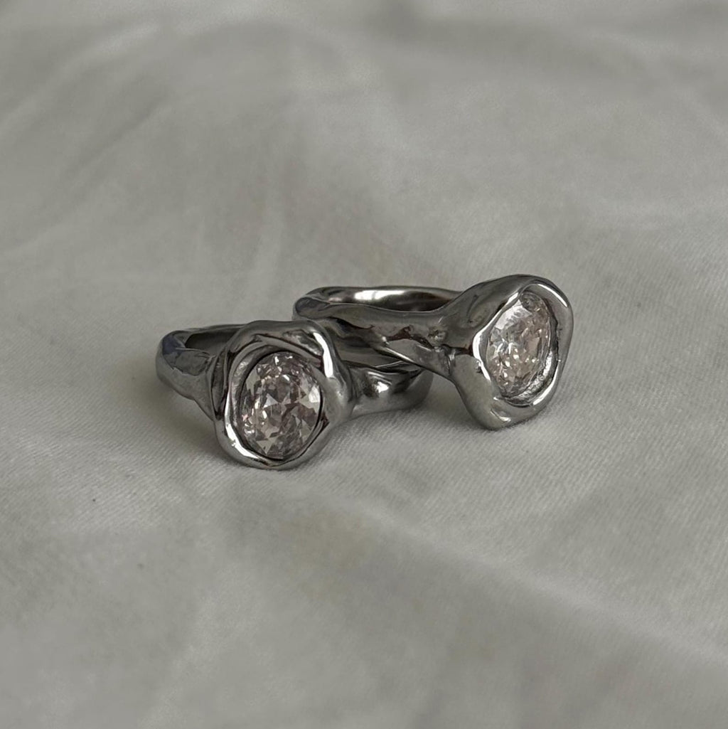 Two silver rings with textured, organic-shaped bezel settings holding clear round gemstones, displayed on a soft, light gray fabric background.
