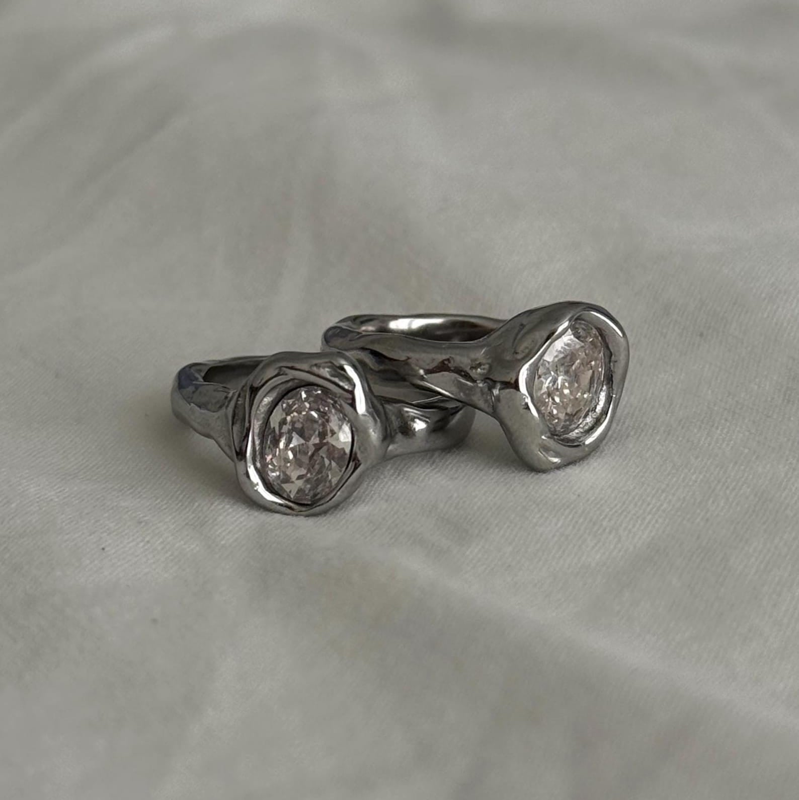 Two silver rings with textured, organic-shaped bezel settings holding clear round gemstones, displayed on a soft, light gray fabric background.