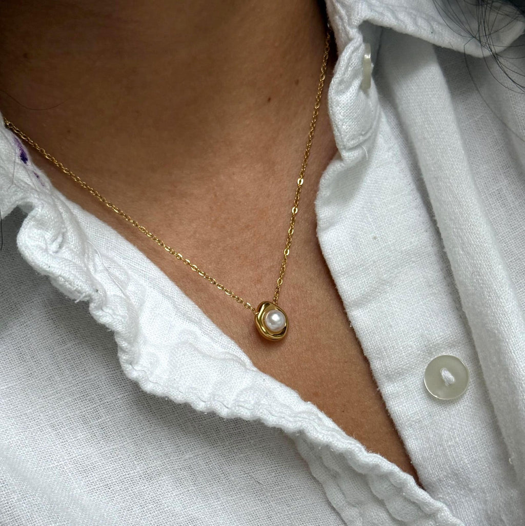 A close-up of a person wearing a delicate gold chain necklace with a small oval pearl pendant, styled with a white textured linen button-up shirt.