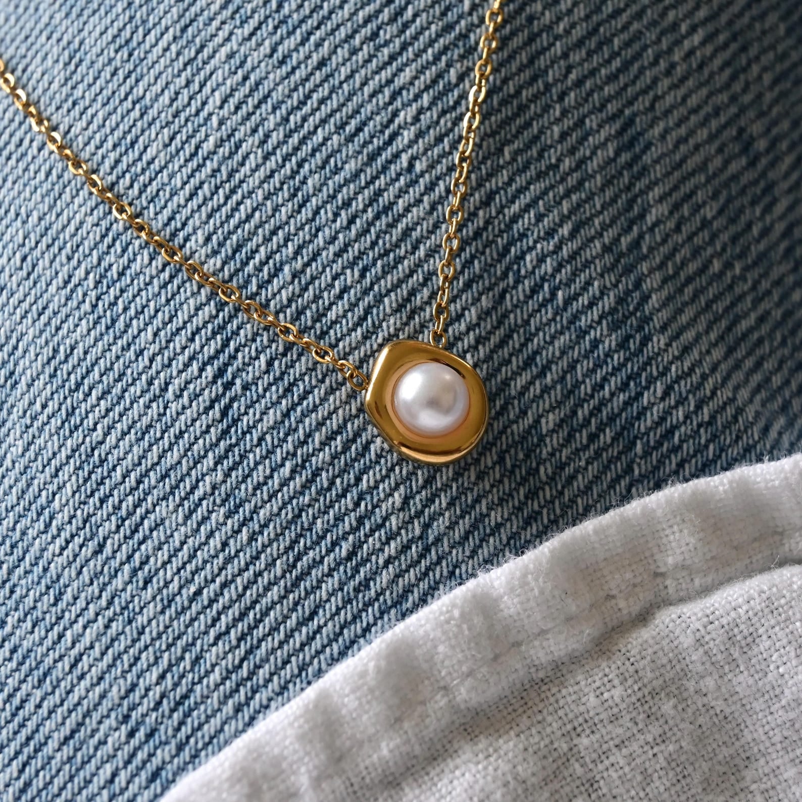 A gold chain necklace with a small gold bezel holding a white pearl pendant, displayed on a blue denim fabric background with a light gray fabric fold at the bottom right.