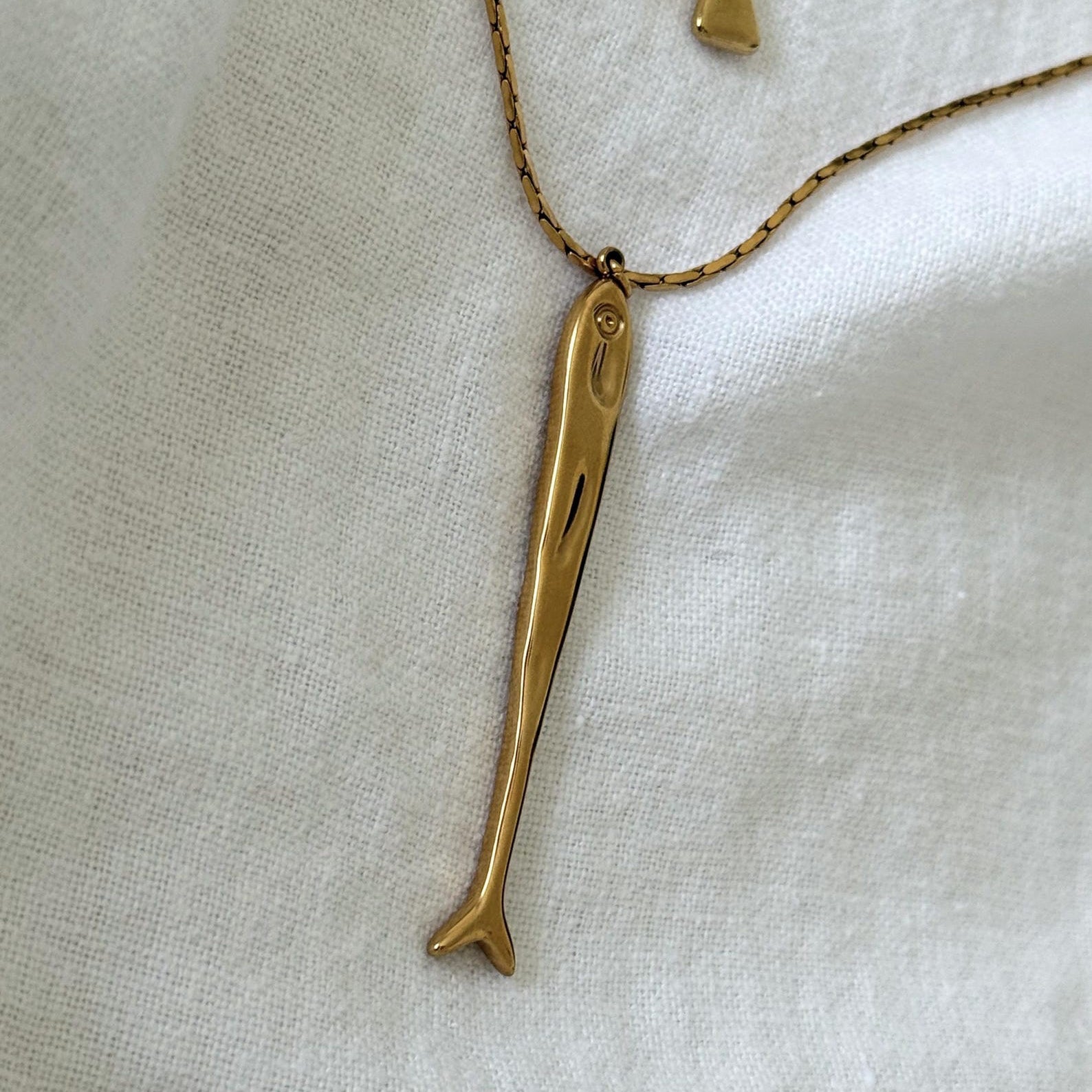 A gold sardine pendant necklace on a delicate gold chain, displayed on a textured white fabric background.