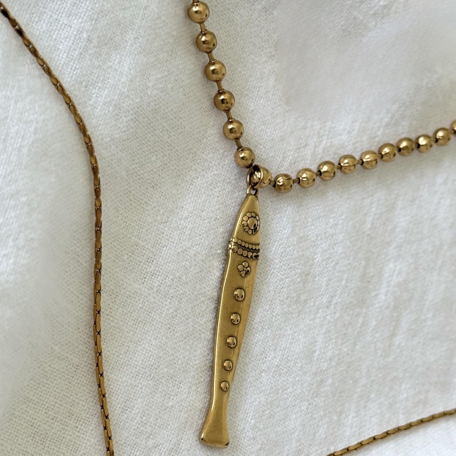 A gold-toned necklace featuring a fish pendant with small bead details, displayed on a textured white fabric background.