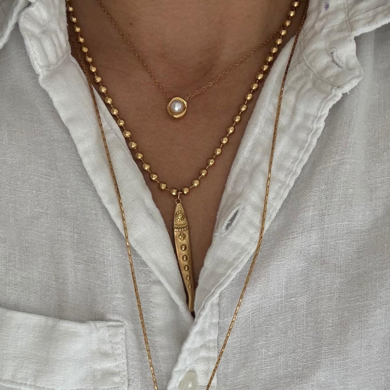 A close-up of a person wearing layered gold necklaces over a white linen button-up shirt. The necklaces include a beaded chain with a fish pendant.