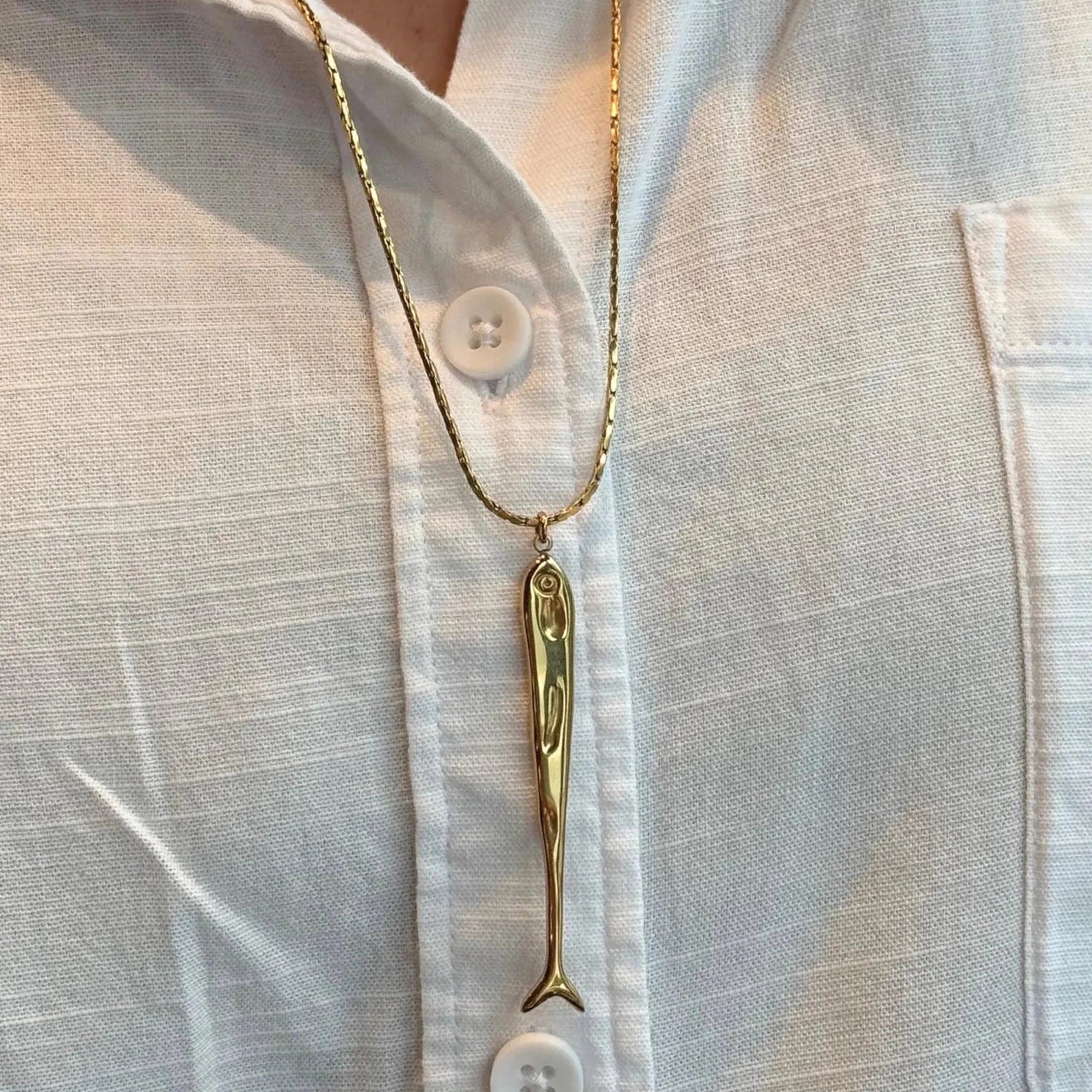 A gold sardine pendant necklace worn on a white button-down shirt. The necklace features a detailed gold fish charm hanging from a delicate gold chain, styled on a model's neck.