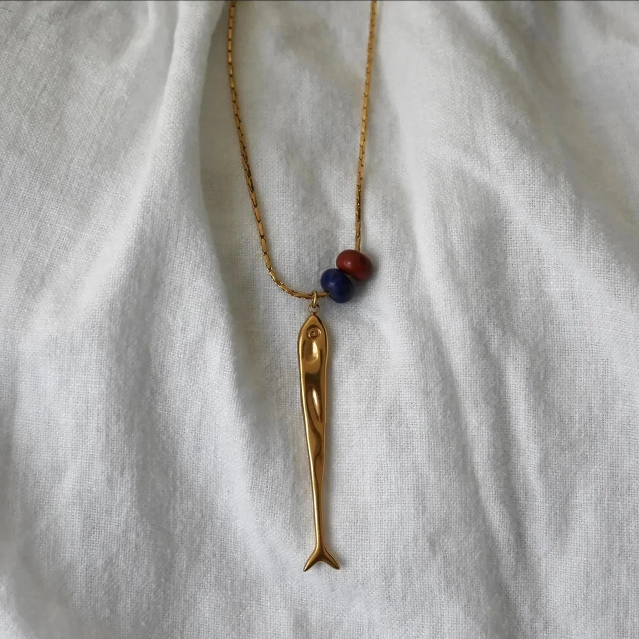 A gold necklace with a stylized fish pendant and two small round beads in dark blue and red, representing Venezuelan flag, laid flat on a white textured fabric background.
