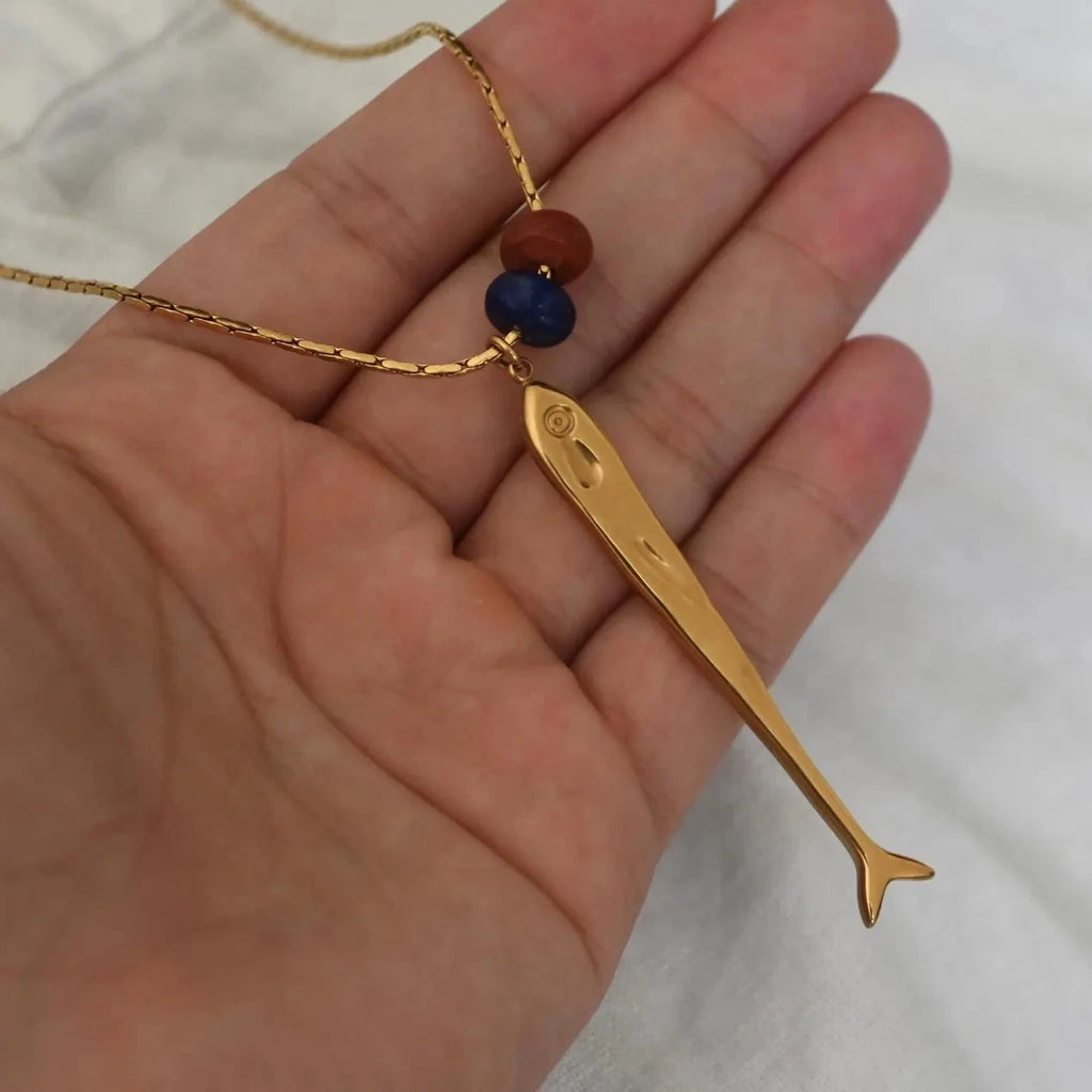 A gold fish pendant necklace with two small beads (one blue and one red) on a gold chain, held in a person's hand against a white fabric background.