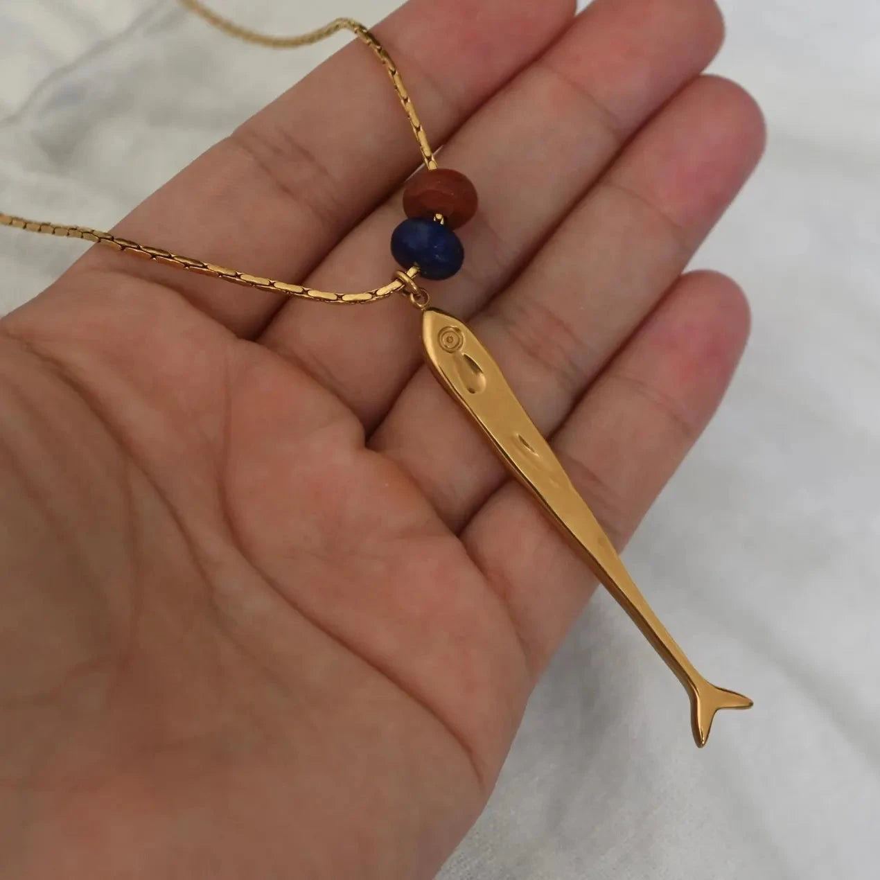 A gold fish pendant necklace with two small beads (one blue and one red) on a gold chain, held in a person's hand against a white fabric background.