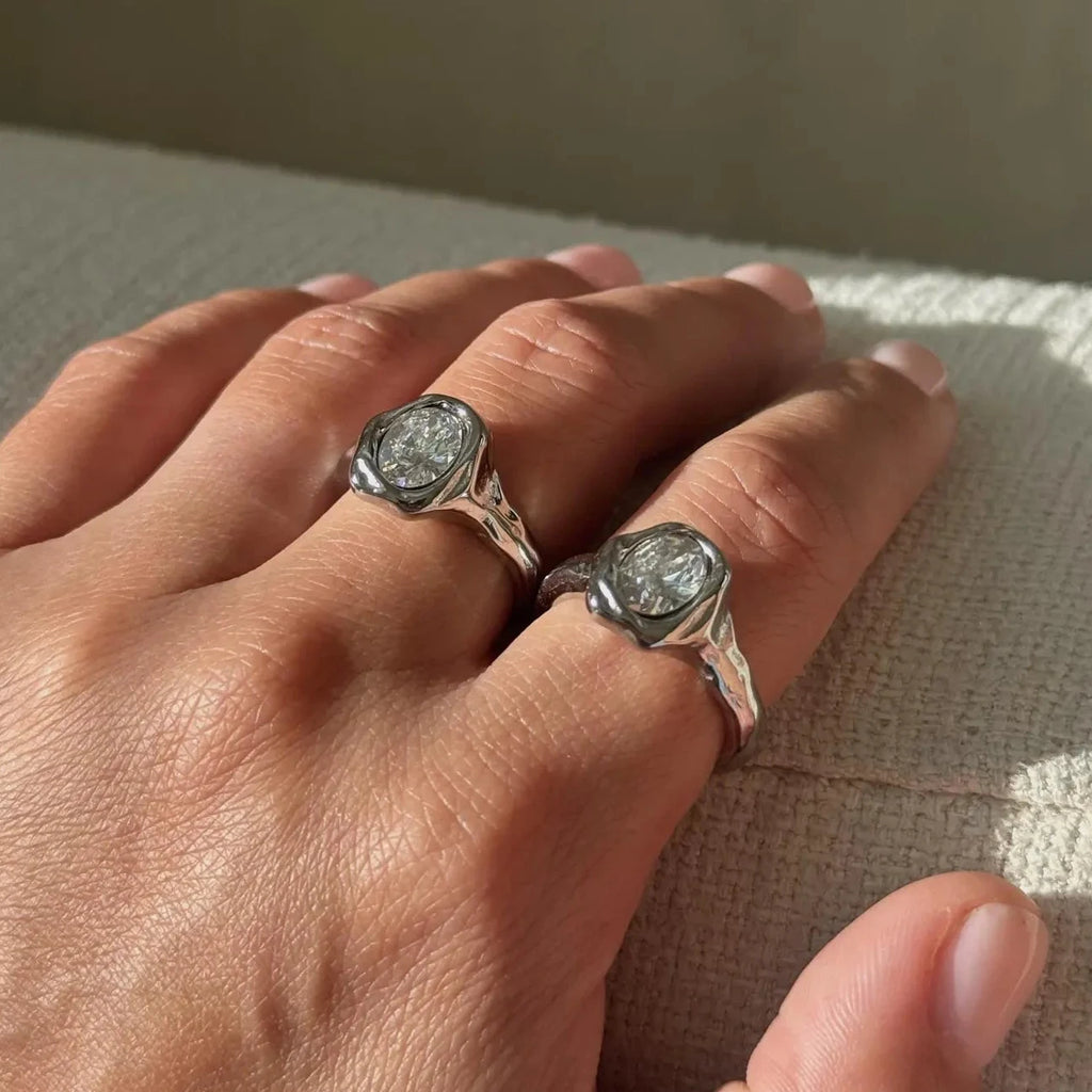 A close-up of a hand wearing two silver molten rings with round bezel-set clear stones, resting on a textured light beige surface.