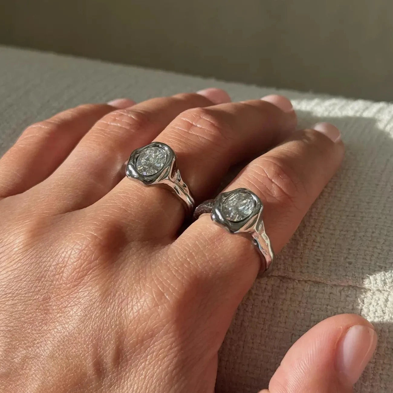 A close-up of a hand wearing two silver molten rings with round bezel-set clear stones, resting on a textured light beige surface.