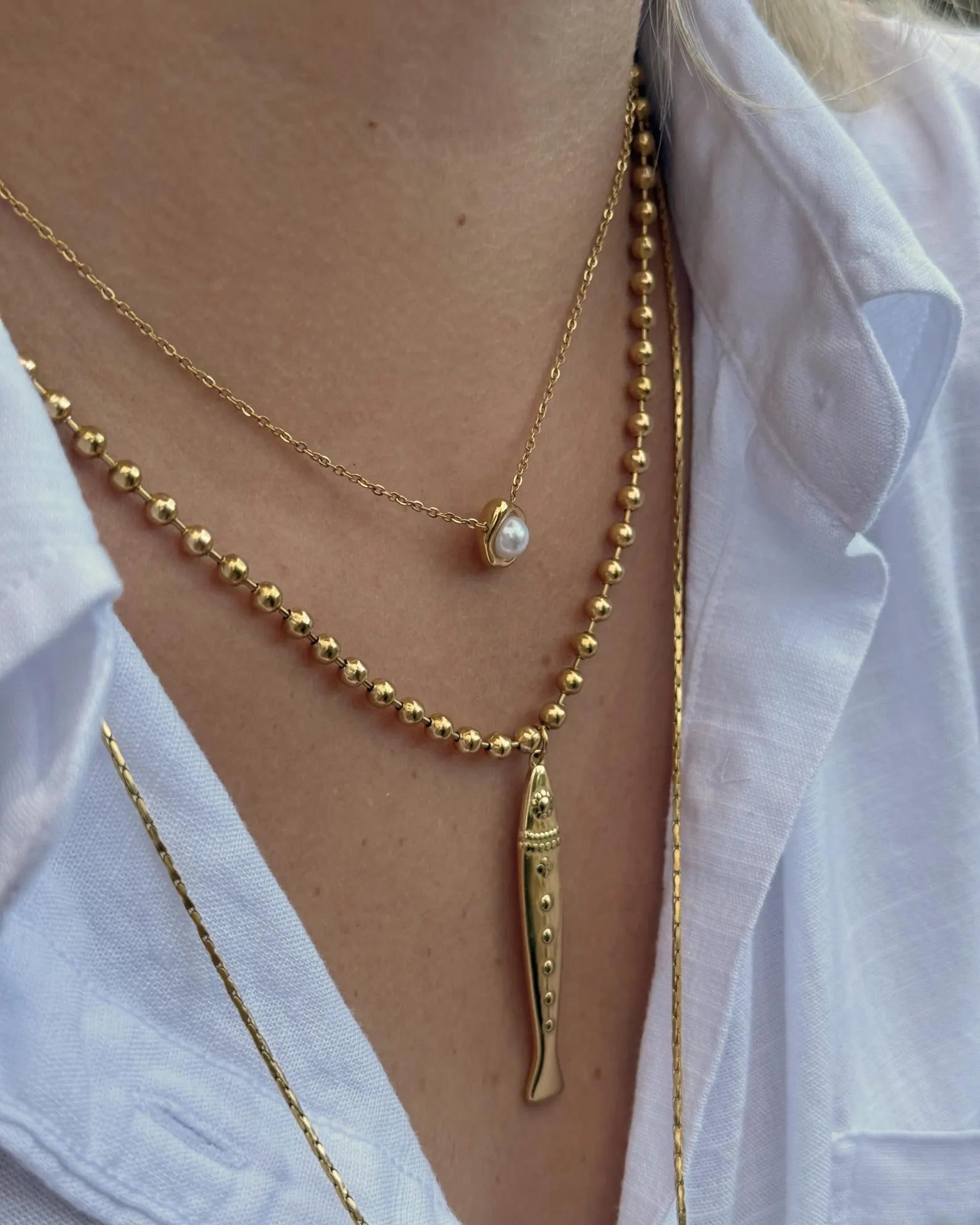 A close-up of a person's neck and chest wearing layered gold necklaces over a white collared shirt. The necklaces include a delicate chain with a small white pearl bead and a longer chain with a gold fish-shaped pendant.
