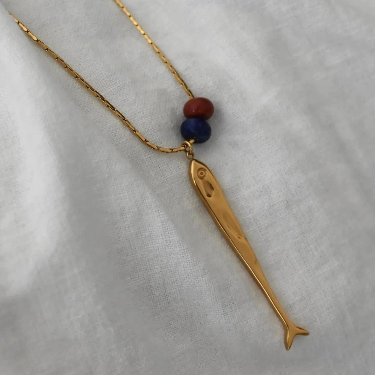 A gold sardine pendant necklace with two small beads (one red, one blue) on a textured white fabric background.
