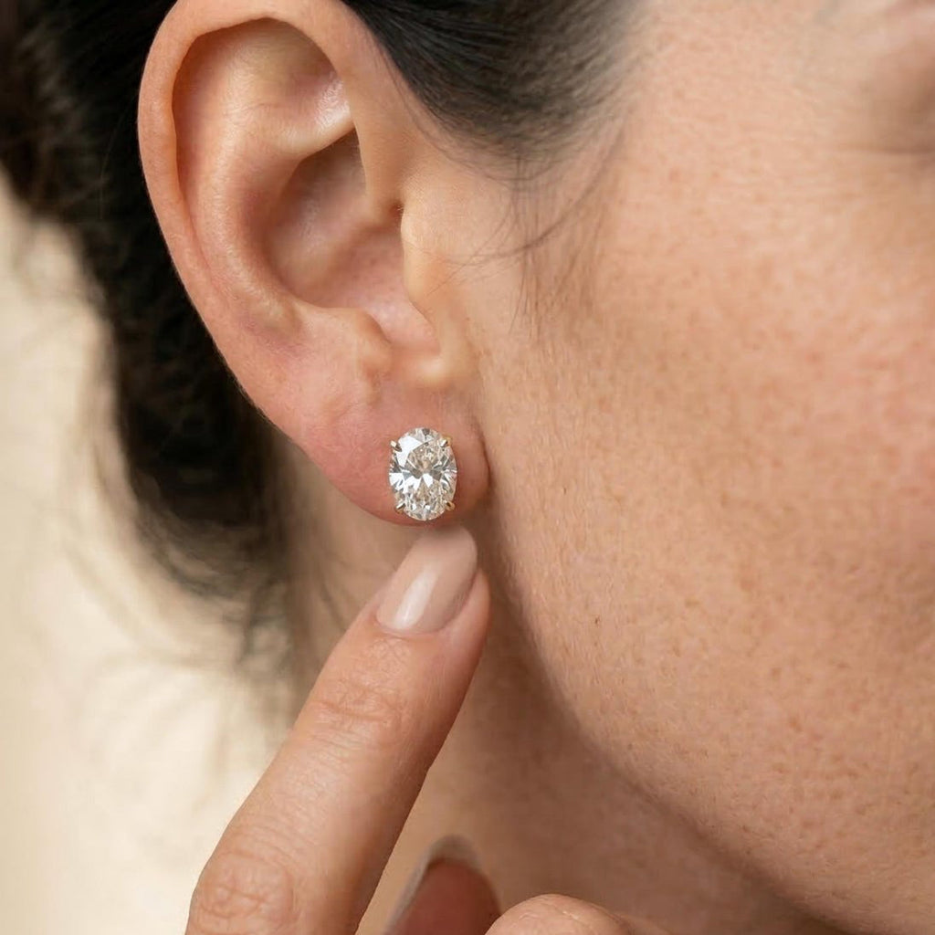 A close-up of a woman wearing a small oval diamond stud earring, with her hand near her ear against a soft beige background.