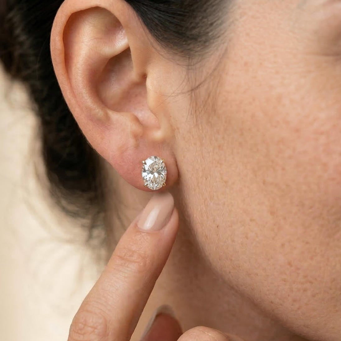 A close-up of a woman wearing a small oval diamond stud earring, with her hand near her ear against a soft beige background.