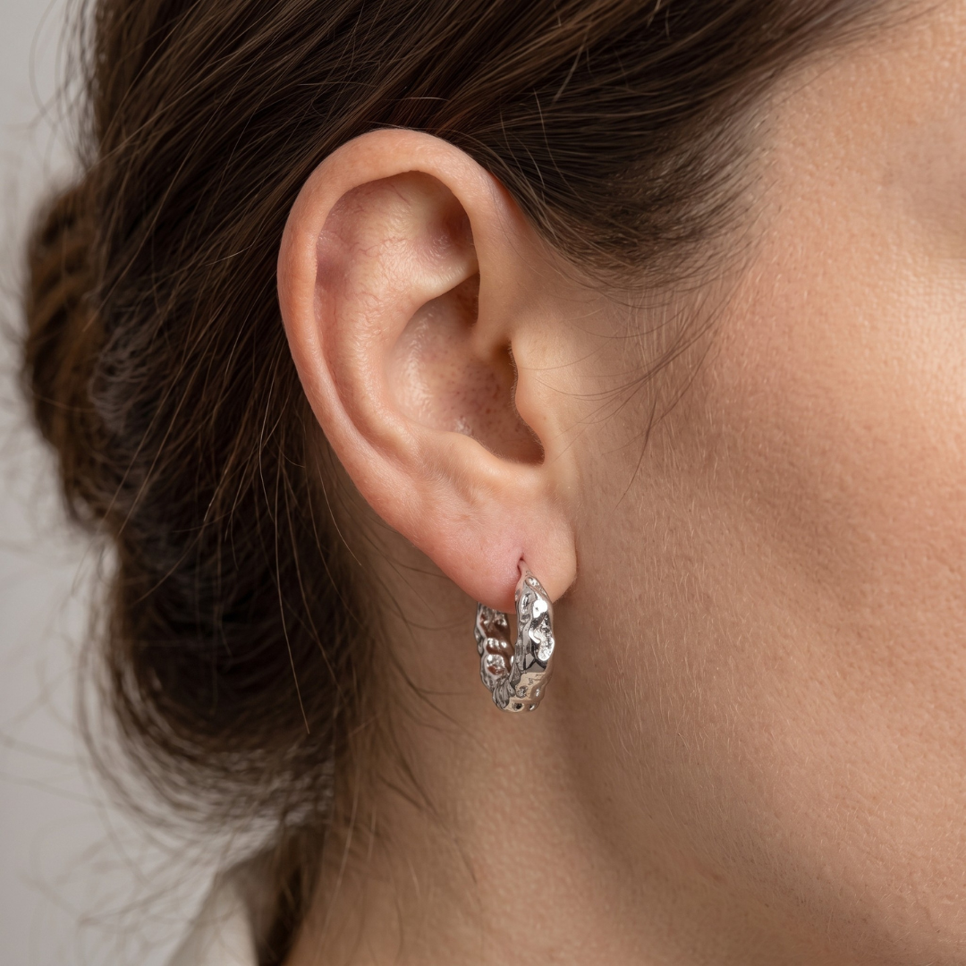 A close-up of a model's ear wearing a small silver huggie hoop earring with textured, organic-shaped metalwork.